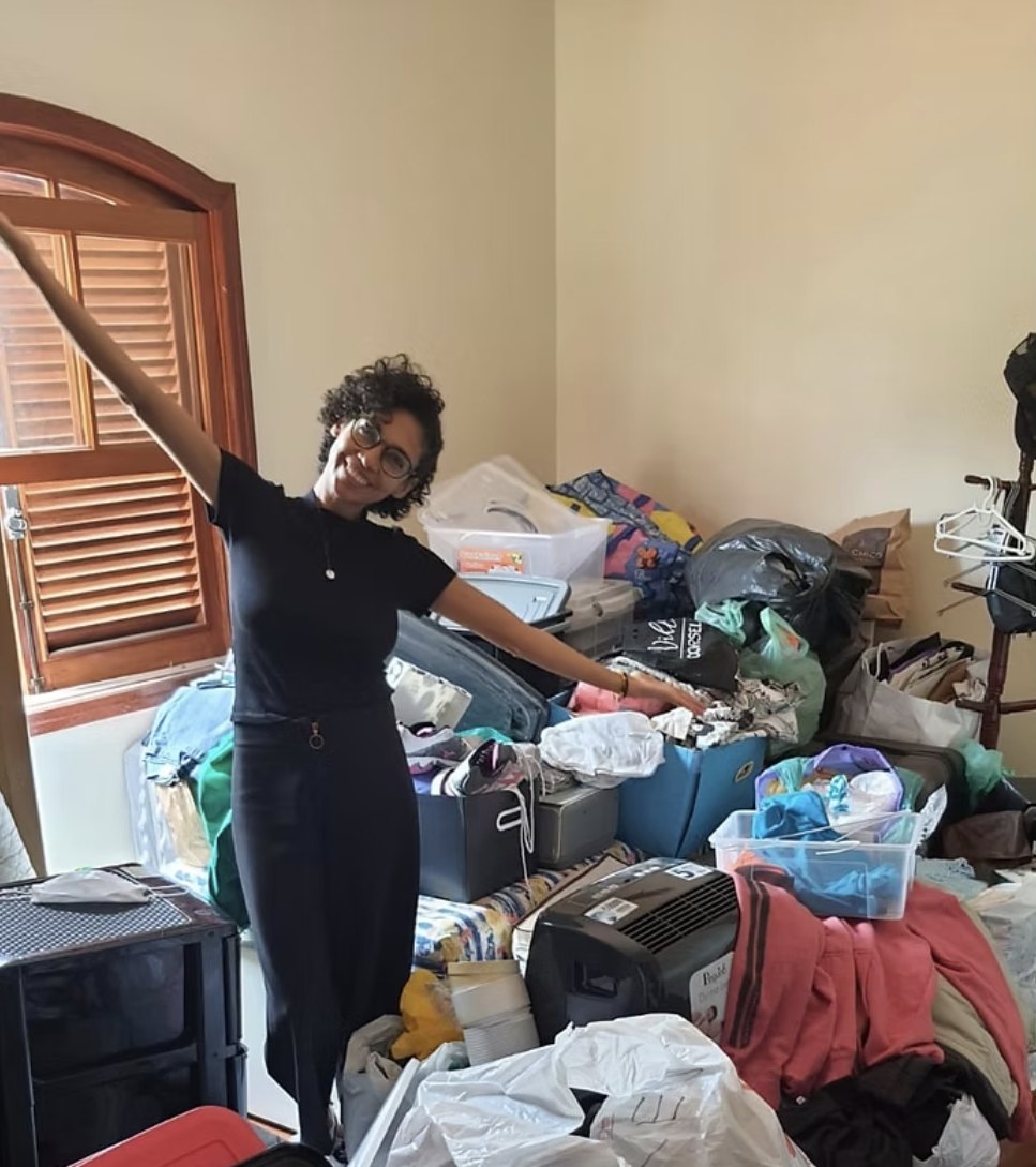 Sheila sorrindo, de braços abertos, em um quarto cheio de objetos prestes a ser organizado
